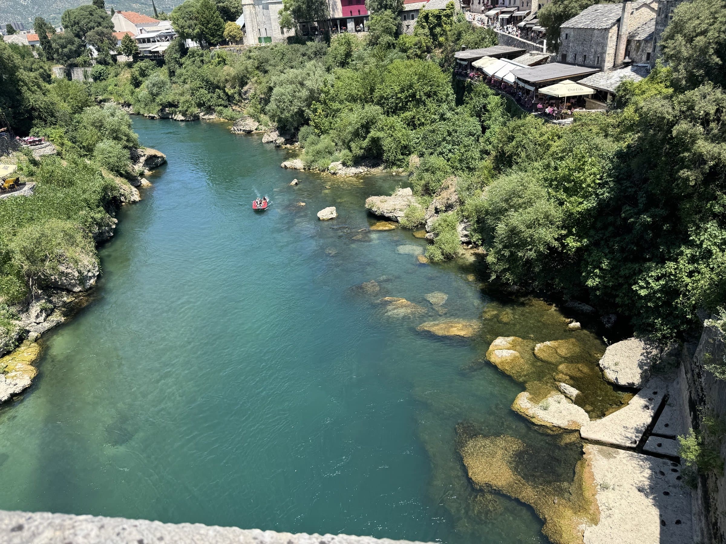 The emerald-green Neretva seen from the Old Bridge in Mostar, a red raft drifting over the pale rocky bed with bazaar terraces on the bank
