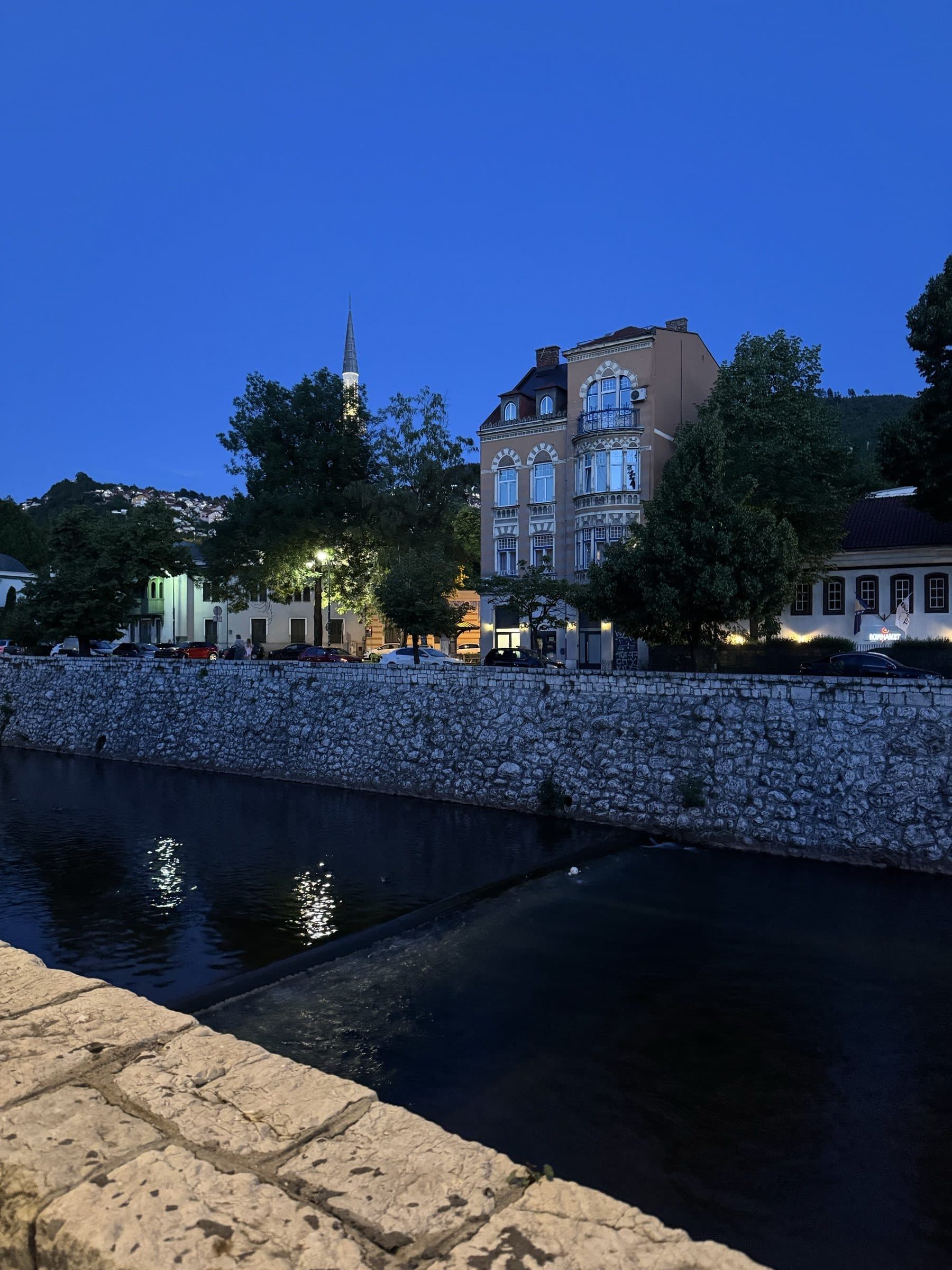 Blue-hour view of the Miljacka river in Sarajevo, streetlamps reflected in the dark water below stone embankment walls, a minaret and old façades against a deep blue sky