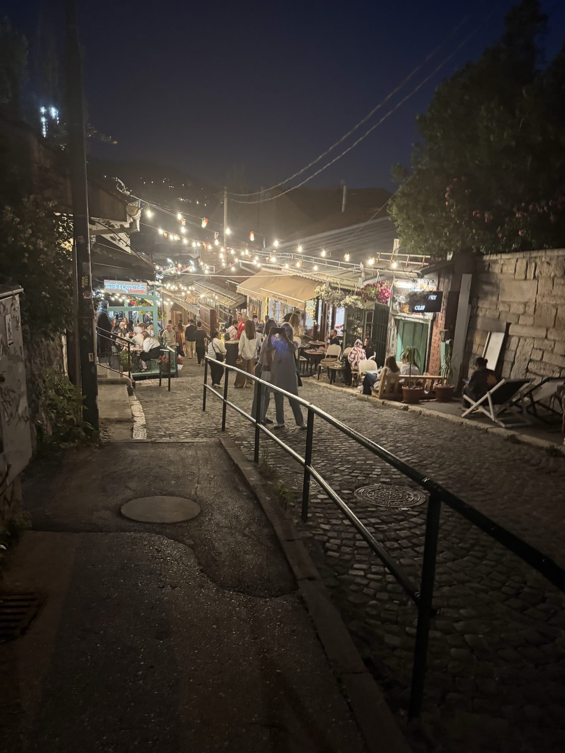 A string-lit cobbled lane climbing through Sarajevo's old town after dark, people gathered at low tables outside small kitchens